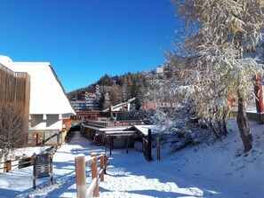A snow-covered outdoor area with a wooden fence, a building with a sloped roof, and a tree with snow on its branches. - Cozy apartment with balcony and pool (Les Arcs)