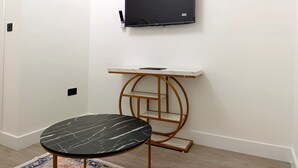 Una habitació amb una paret blanca, un televisor pla fixat a la paret, una taula rodona negra amb un dibuix que imita el marbre, i un moble prestatgeria blanc amb motius de marbre i una estructura de fusta.