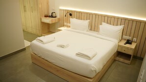 A neatly made bed with white linens and towels, a wooden headboard, a bedside table with a lamp, and a small shelf with a kettle and a box. - Vox Holiday (Palakkad)