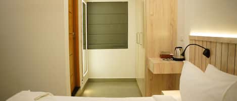 A hotel room with a bed, a bedside table with a lamp, a wooden headboard, a small wooden shelf with a kettle, and a green sliding door.