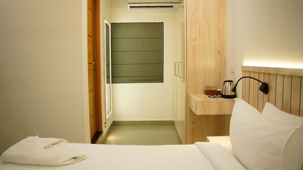 A hotel room with a bed, a bedside table with a lamp, a wooden headboard, a small wooden shelf with a kettle, and a green sliding door. - Vox Holiday (Palakkad)