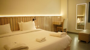 A hotel room with a large bed, a wooden headboard, a bedside table, a chair, a small desk, and a mirror. - Vox Holiday (Palakkad)