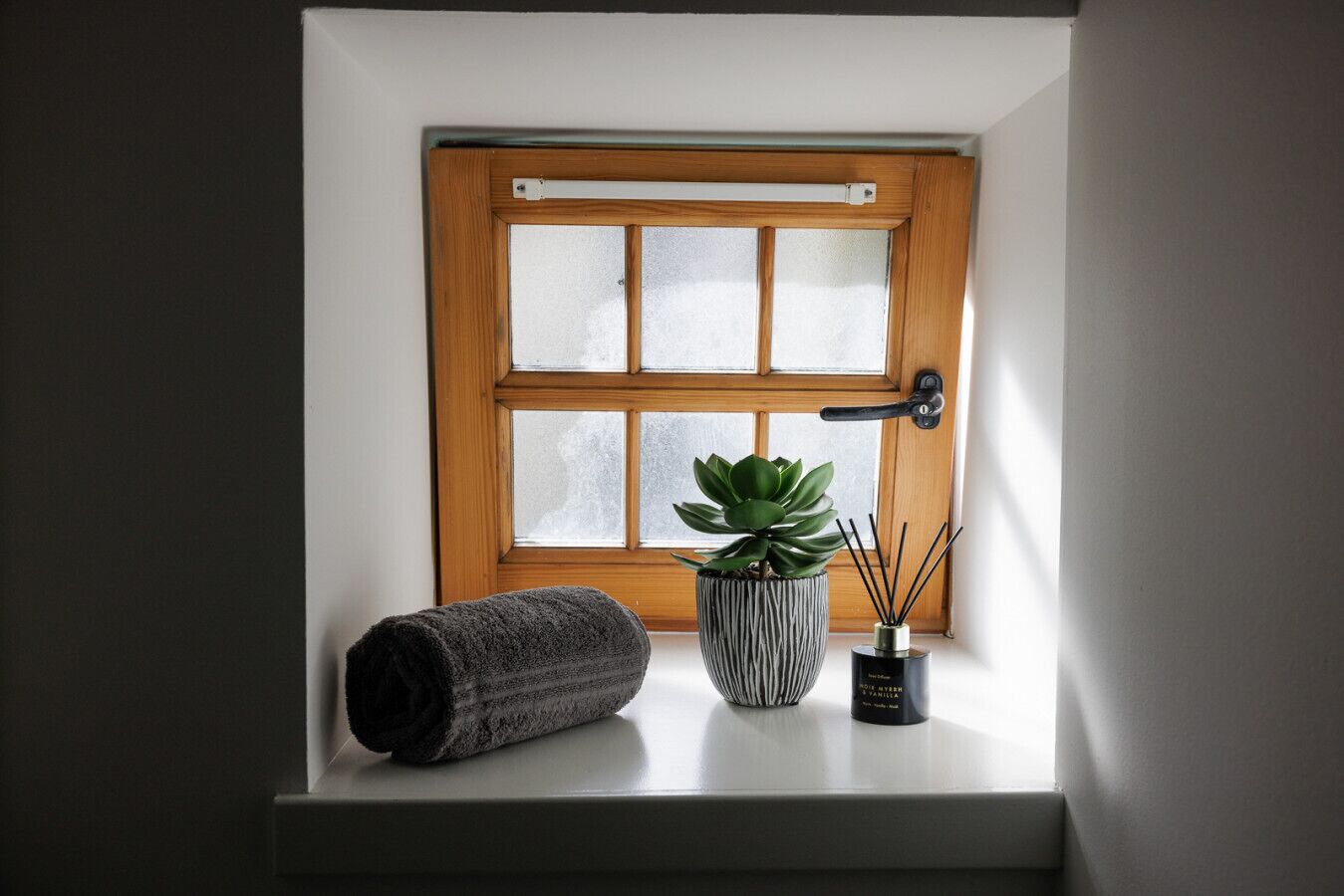 A window with a wooden frame, a rolled towel, a potted plant, and a container with reed sticks.