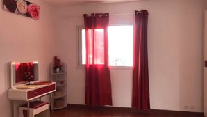Una habitación con piso de madera, un escritorio blanco con una silla roja, un espejo y una ventana con cortinas rojas.