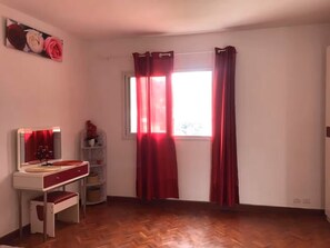 A room with a wooden floor, a white desk with a red chair, a mirror, and a window with red curtains.