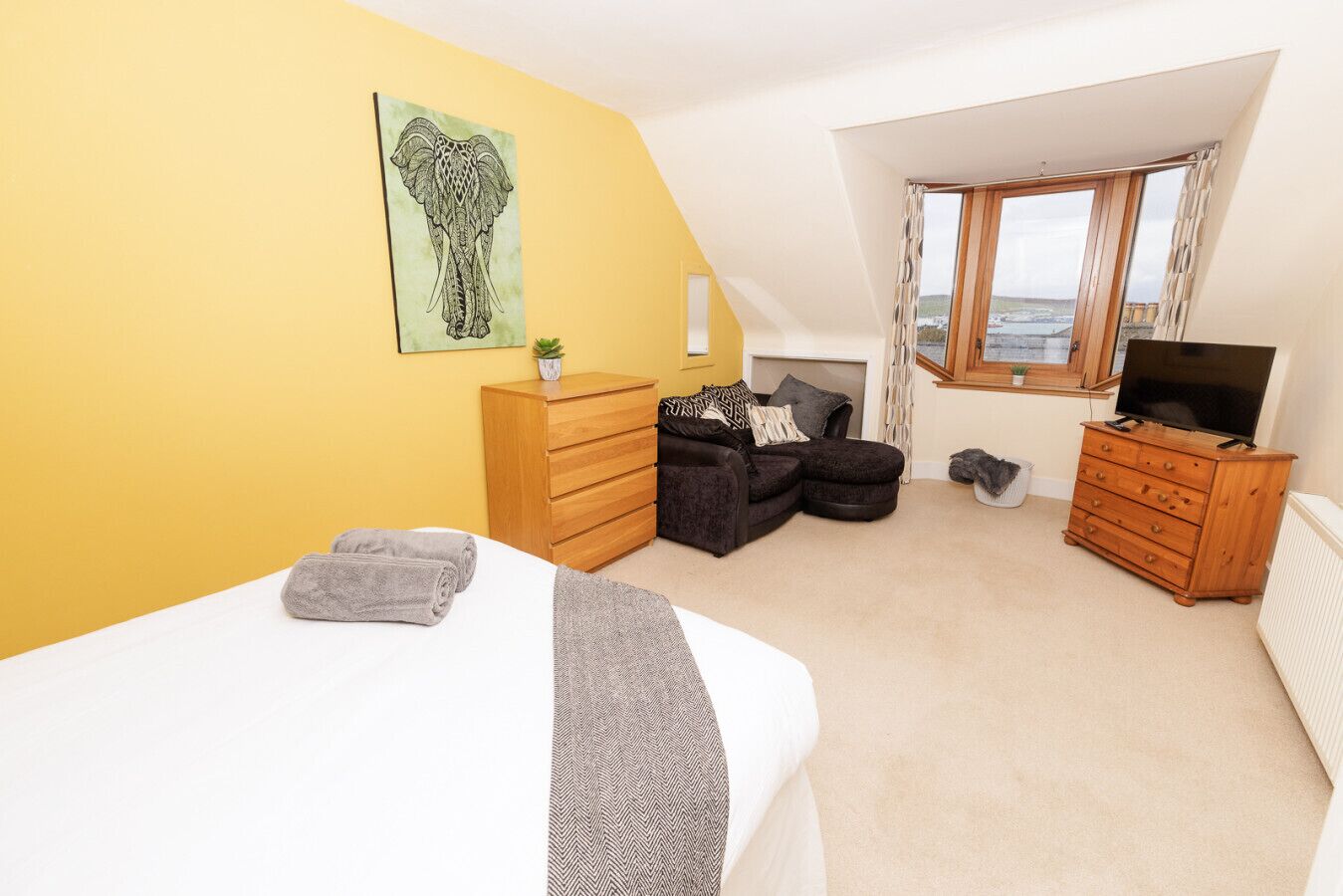 A bedroom with a bed, a wooden dresser, a black sofa, a television, and a window with a view.