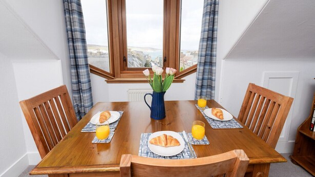 A dining table with four chairs, a vase with flowers, and plates with croissants and orange juice.