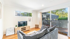 Een moderne woonkamer met een flatscreen-tv, een houten tv-meubel, een grijze bank en een balkon met uitzicht op groen.