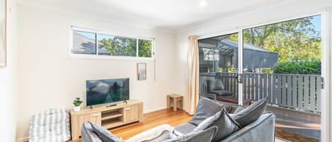 A modern living room with a flat-screen TV, a wooden entertainment unit, a grey sofa, and a balcony with a view of greenery.