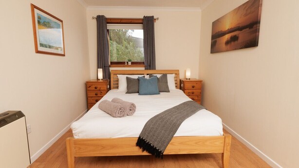 A neatly made bed with a wooden frame, two bedside tables with lamps, a window with curtains, and a painting on the wall. - NEW to VRBO: Peaceful Lerwick Stay; Large Garden, Isles Views (Lerwick)