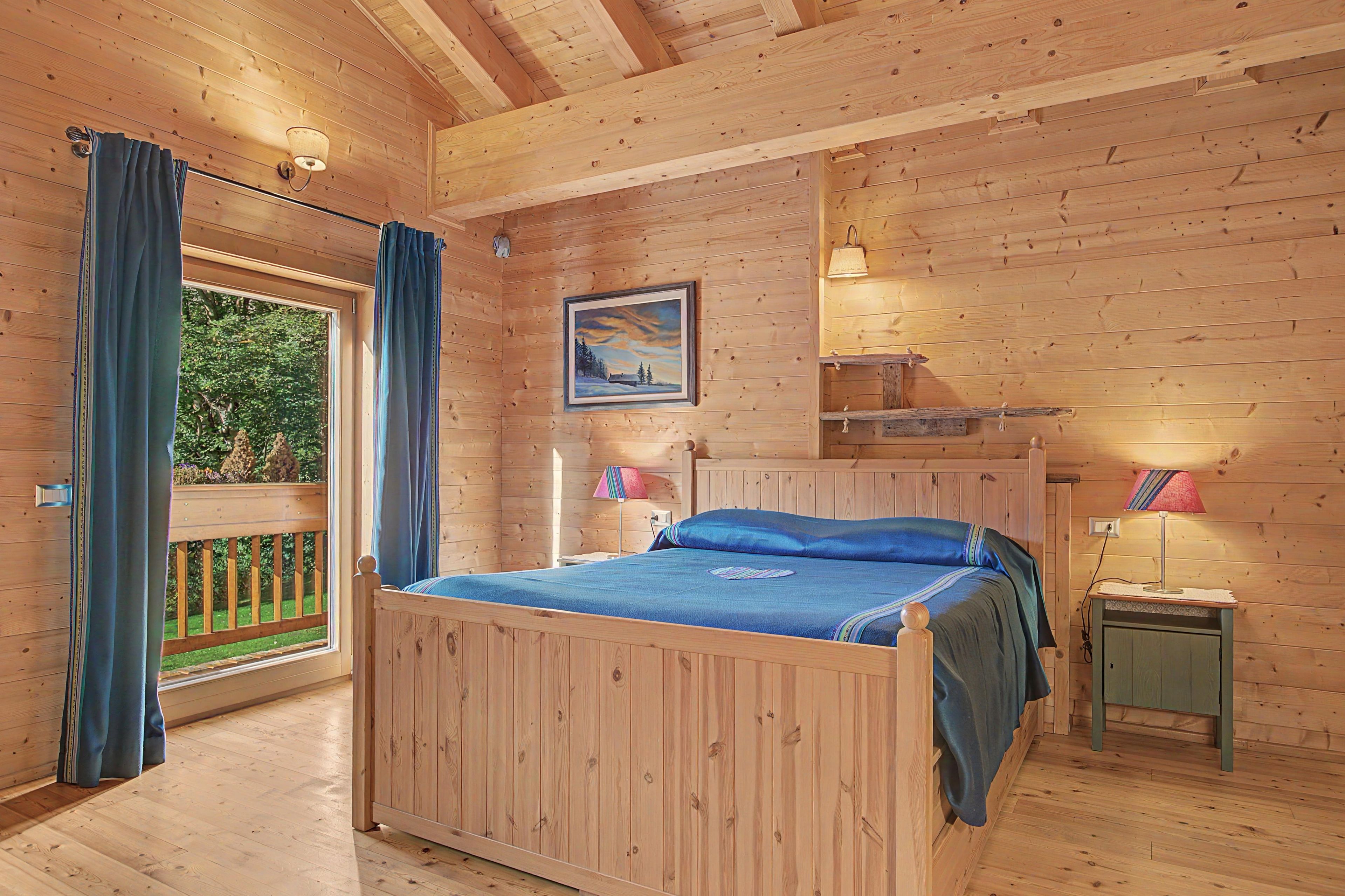 A wooden cabin bedroom with a bed, two bedside lamps, a painting, and a window with curtains.