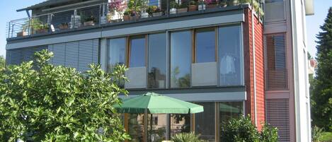 A modern two-story building with a balcony, a green umbrella, and potted plants.