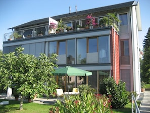 Een modern, tweelaags gebouw met balkon, een groene parasol en potplanten.