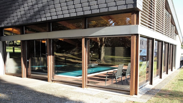 Una casa moderna con un patio con paredes de vidrio, una piscina y mobiliario exterior.