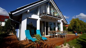 Une maison moderne à deux étages, dotée d’un balcon, d’une terrasse en bois et d’un espace salon extérieur.