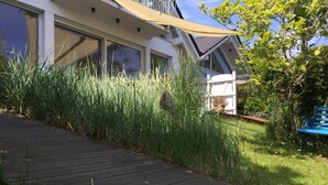 Une terrasse en bois mène à une maison dotée de grandes fenêtres, d’une clôture blanche et d’un banc bleu dans le jardin.