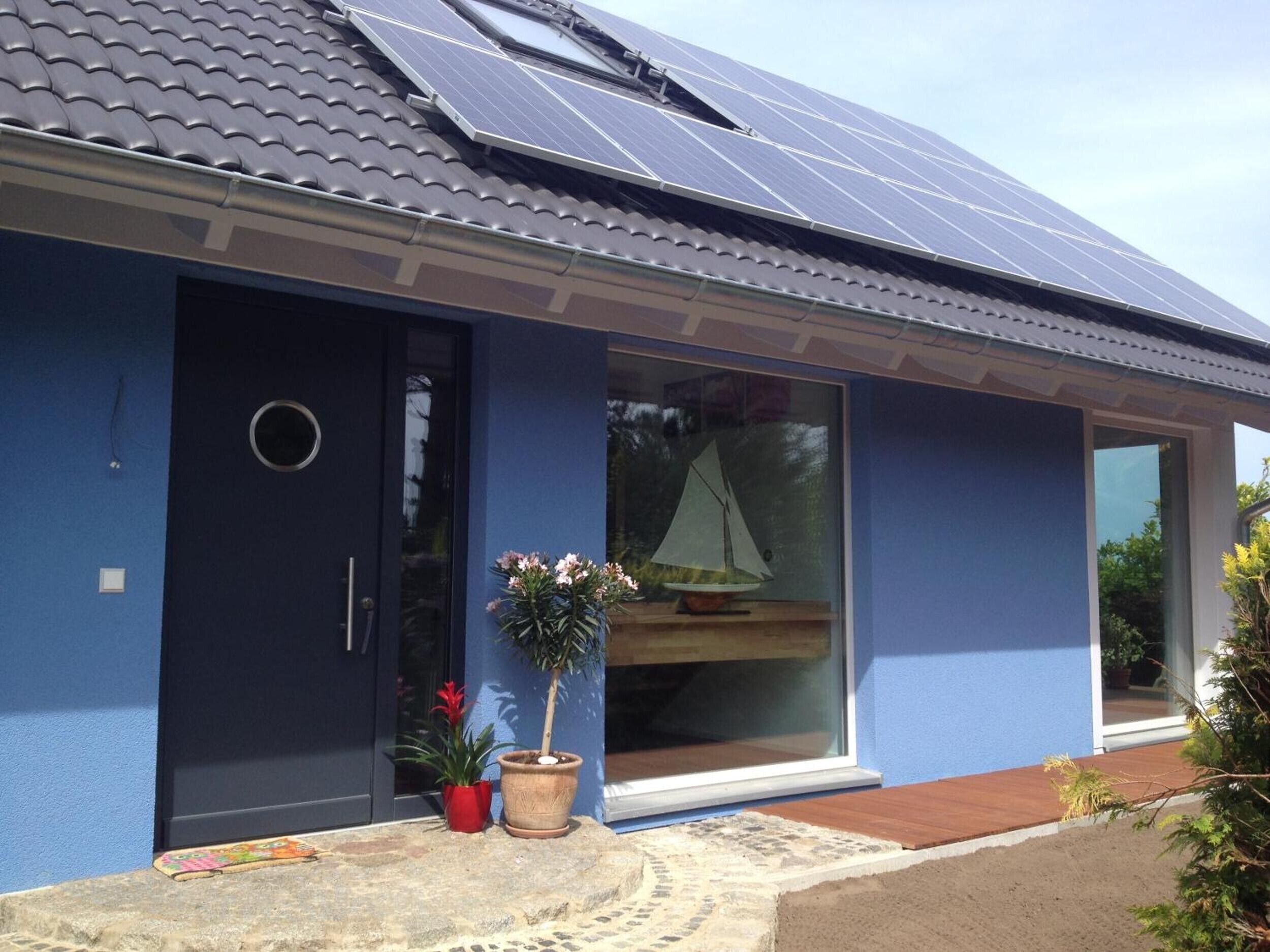 Una casa con paneles solares, fachada azul oscuro, puerta corredera de vidrio y un pequeño jardín con plantas en macetas.