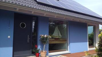 Una casa con paneles solares, fachada azul oscuro, puerta corredera de vidrio y un pequeño jardín con plantas en macetas.