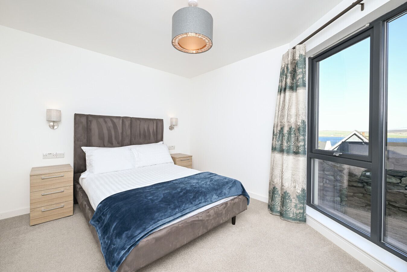 A modern bedroom with a large bed, bedside tables, a lamp, and a view of a building through a window.