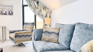 A modern living room with a grey sofa, a patterned armchair, and a large window with curtains. - NEW to VRBO: Elegant Lerwick Townhouse • Harbour Views + Patio (Lerwick)