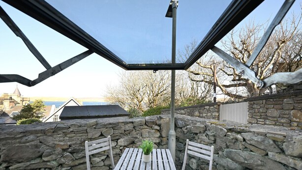A stone wall with a wooden table and two chairs, a metal pole, and a view of buildings and trees. - NEW to VRBO: Elegant Lerwick Townhouse • Harbour Views + Patio (Lerwick)