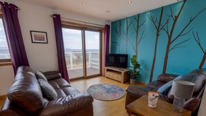 A living room with a brown leather sofa, a wooden coffee table, a television, and a view of the sea through a sliding glass door.