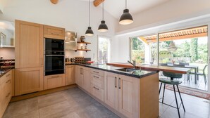 Une cuisine moderne avec des armoires en bois, un four encastré, un grand îlot central doté d’un évier, des luminaires suspendus et une vue sur l’extérieur.