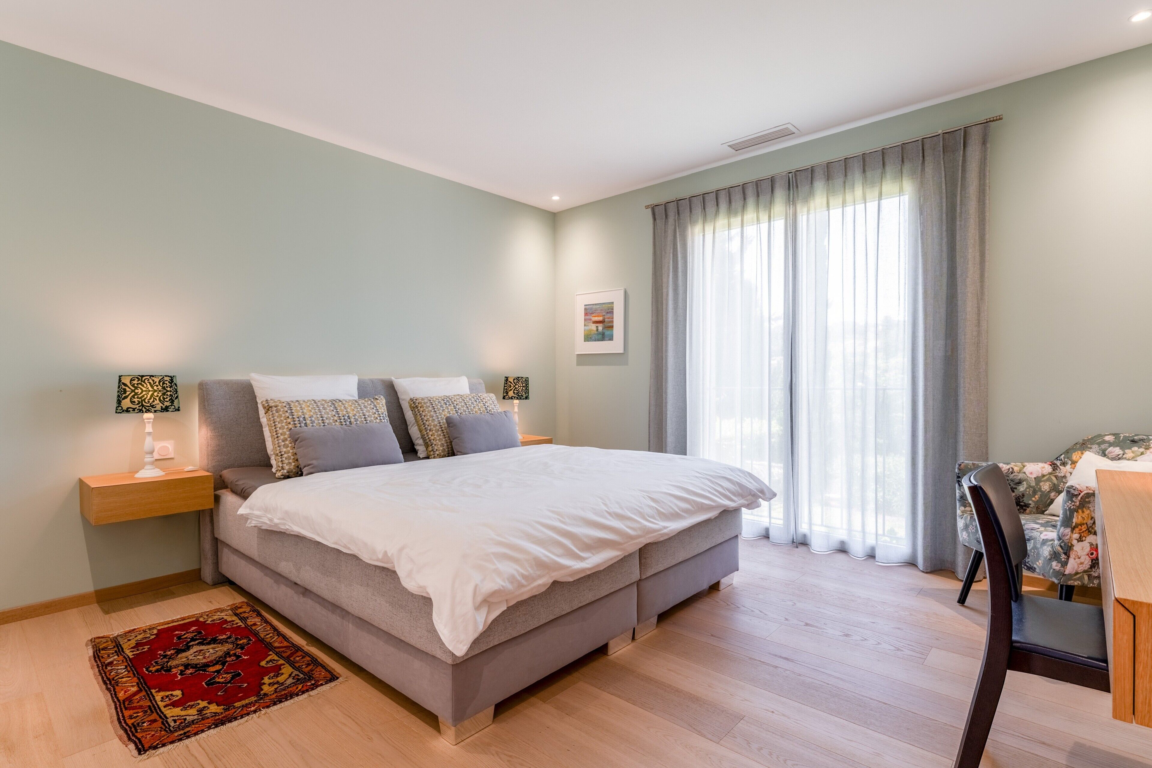 Un dormitorio con cama, mesitas de noche, alfombra, silla y ventana con cortinas.