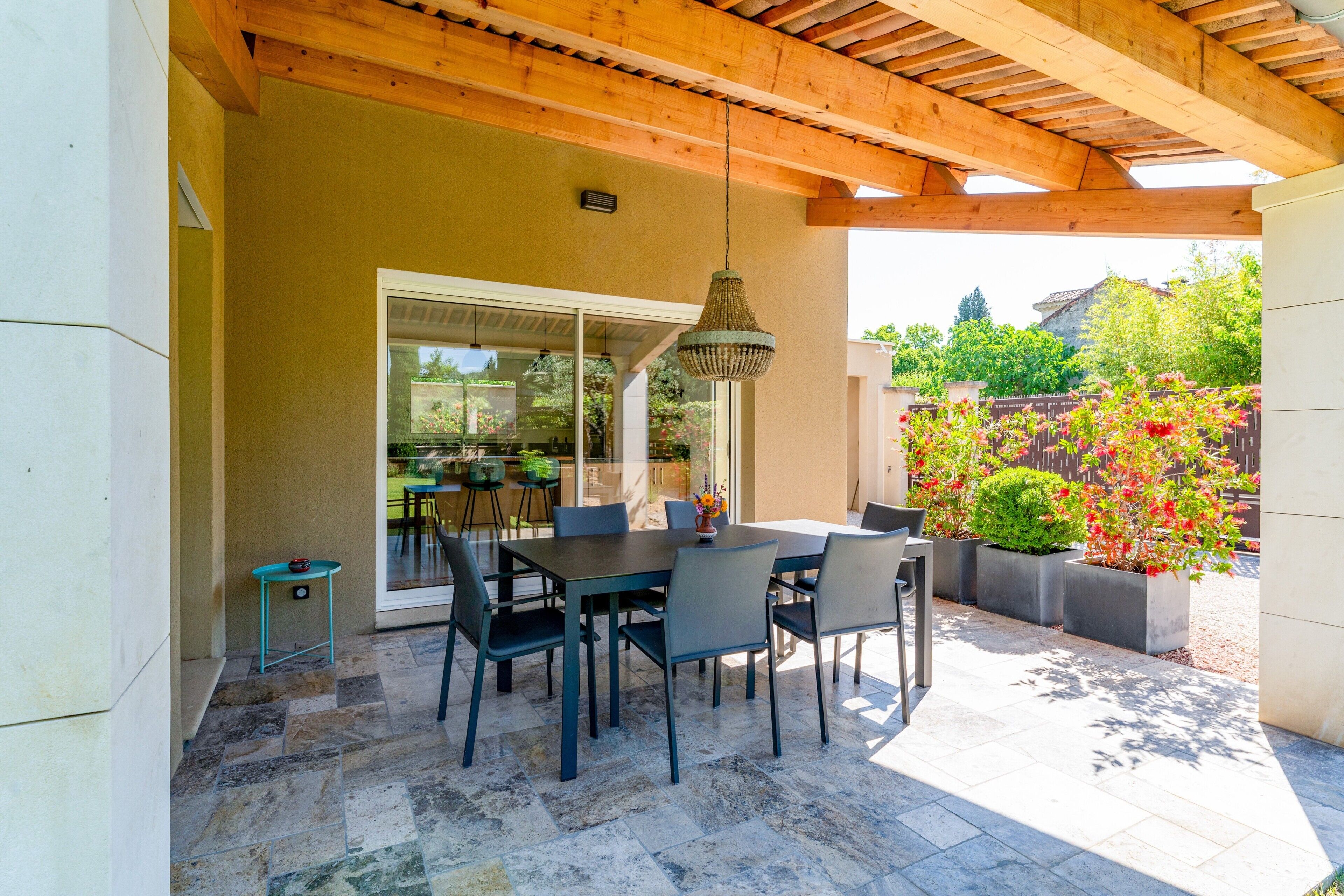Une terrasse extérieure avec une table et des chaises, un luminaire suspendu et une vue sur un jardin fleuri.