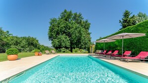 Une piscine rectangulaire à l’eau d’un bleu limpide, entourée de chaises longues rouges et de parasols, avec un grand arbre et une colline verdoyante en arrière-plan.
