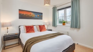 Une chambre à coucher comprenant un lit, deux tables de chevet, une commode, une fenêtre avec des rideaux, un radiateur, un luminaire fixé au mur et un tableau représentant un paysage urbain.