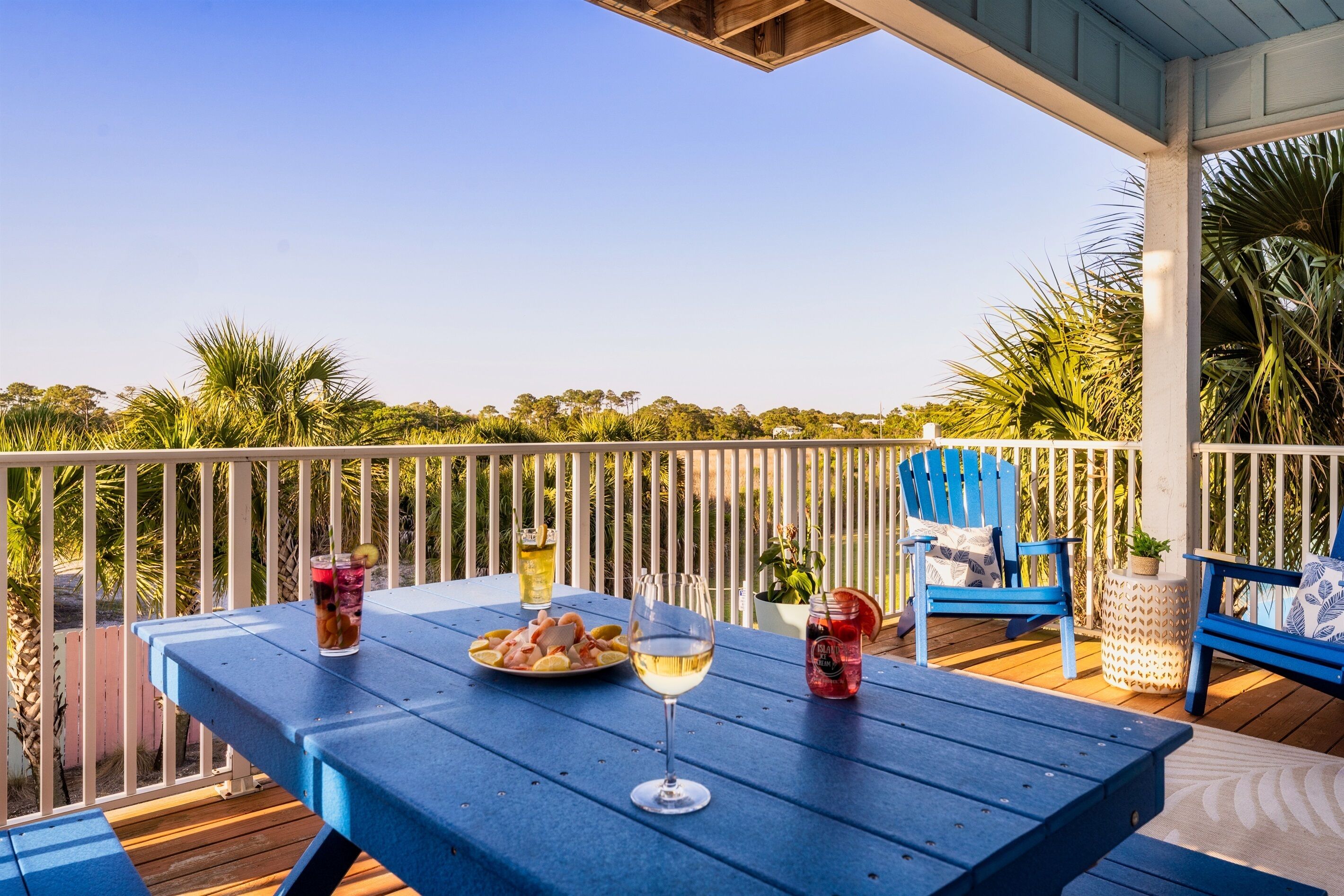 A blue table with drinks and a plate of food on a balcony with white railings and palm trees in the background.