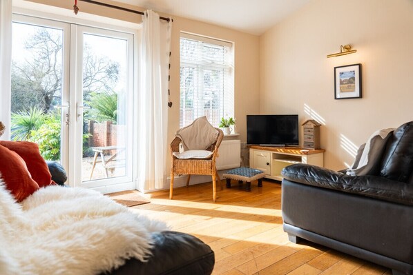 Ein Wohnzimmer mit einer Couch, einem Fernseher, einem Sessel und einem Fenster mit Vorhängen.