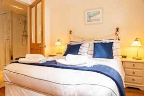 Ein ordentlich bezogenes Bett mit einer blau-weißen Streifenauflage, zwei Kissen und einem weißen Handtuch auf der Seite. Zwei Nachttischlampen, eine Holztür und ein gerahmtes Bild sind an der Wand zu sehen.