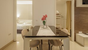 Moderni hotellihuone, jossa on lasinen ruokapöytä, maljakko kukkineen, valkoisilla lakanoilla varustettu sänky ja puusta tehty koristeellinen seinä.