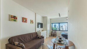A modern living room with a brown sofa, a dining table with a set of glasses and plates, and a city view through a large window. - Studio in Barra 600 m From the Beach Bsf0806 (Salvador)