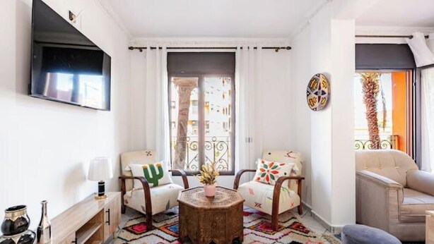 Ein Wohnzimmer mit an der Wand montiertem Fernseher, zwei Sesseln mit dekorativen Kissen, einem runden Holztisch und einem farbenfrohen Teppich.