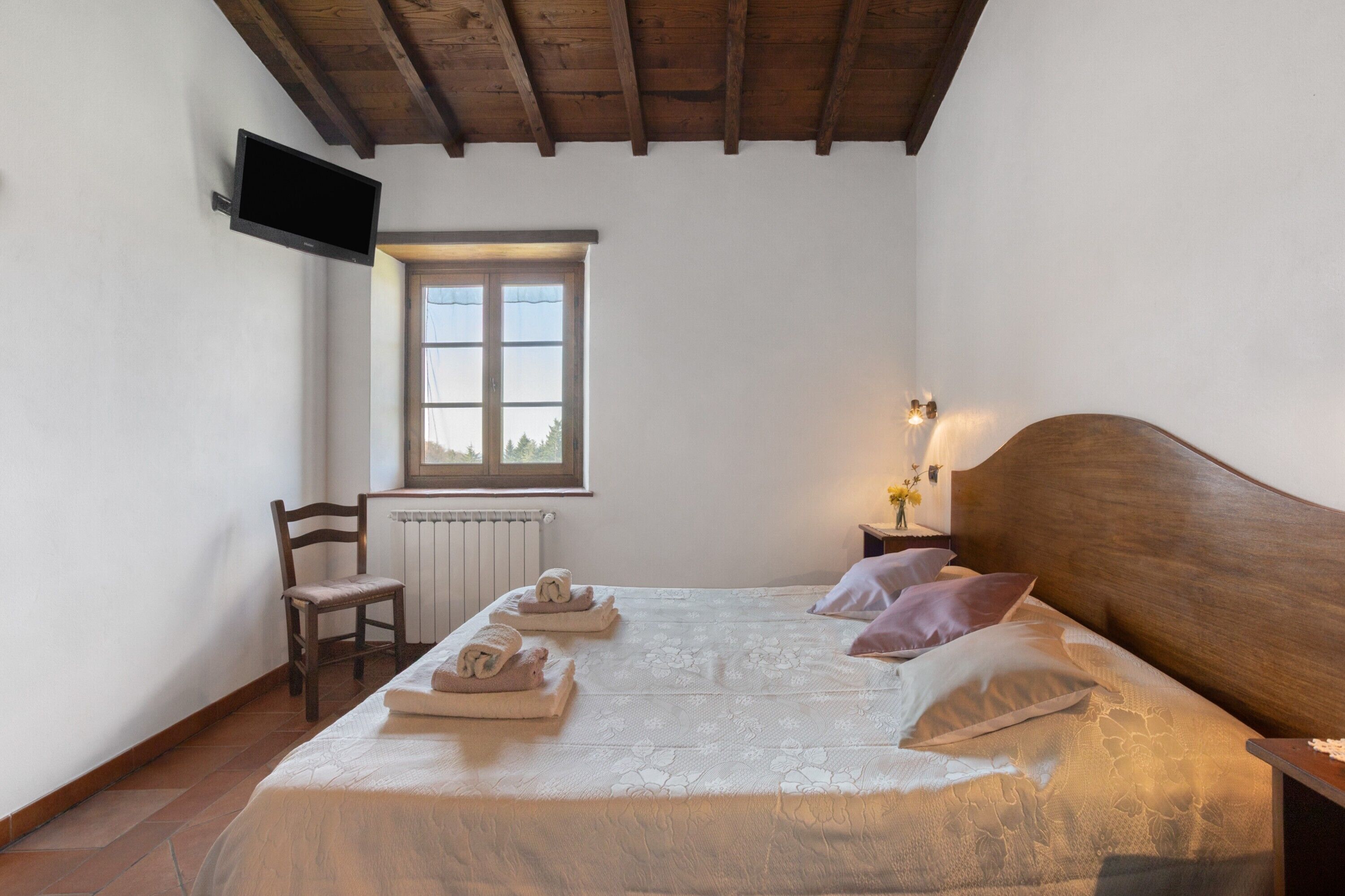 Ein Schlafzimmer mit einem Bett, einem Sessel, einem an der Wand befestigten Fernseher, einem Fenster und einem Nachttisch mit einer Lampe.