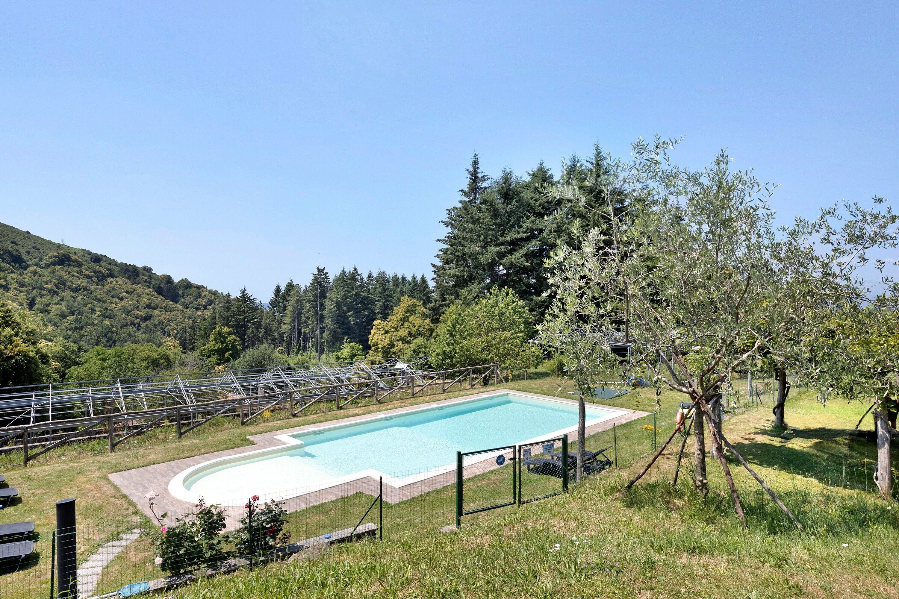 A swimming pool with a surrounding fence, trees, and a hilly landscape in the background.