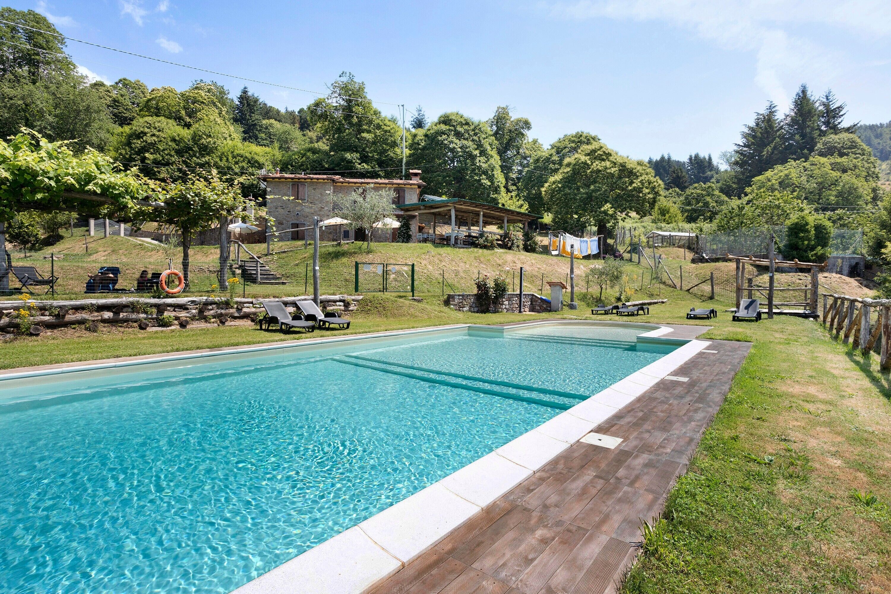 A rectangular swimming pool with a wooden deck, surrounded by a grassy area with lounge chairs and a stone building in the background.