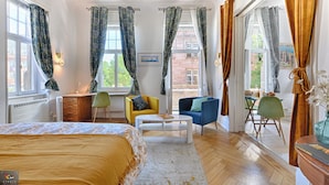 A well-lit room with a bed, a desk, two chairs, a coffee table, and large windows with patterned curtains. - City Suite German Chic The Imperial Balcony (Metz)