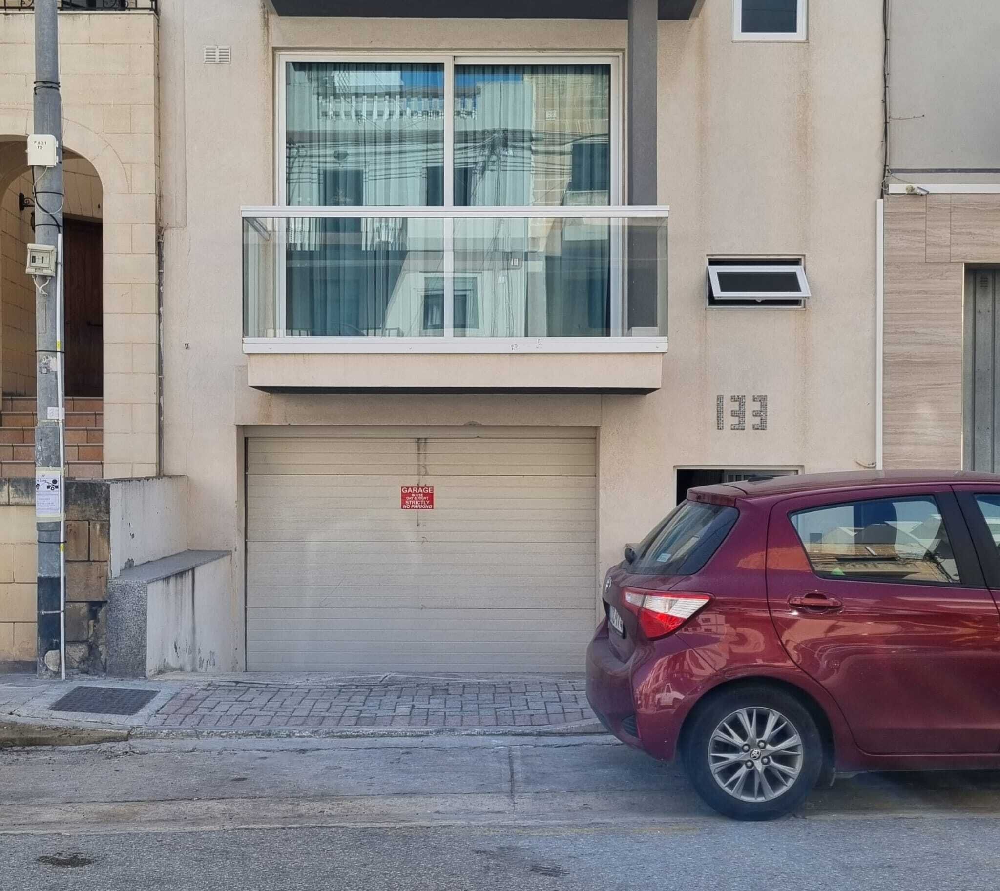 A building with a garage door, a red car parked in front, and a utility pole with a sign.