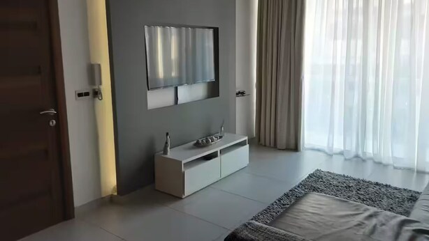 A modern bedroom with a large bed, a TV mounted on the wall, a white console table, and a window with sheer curtains. - Modern 3BR Apartment in Swieqi (Is-Swieqi)