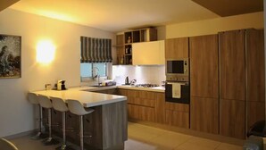 A modern kitchen with a central island, bar stools, wooden cabinets, a microwave, and a stove. - Modern 3BR Apartment in Swieqi (Is-Swieqi)