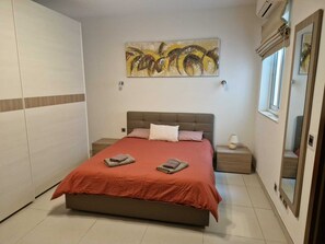 Un dormitorio con cama, mesita de noche, aire acondicionado instalado en la pared y ventana con vistas.