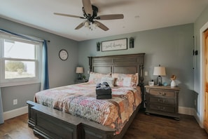 A bedroom with a bed, a nightstand, a ceiling fan, a window with curtains, and a wall clock.