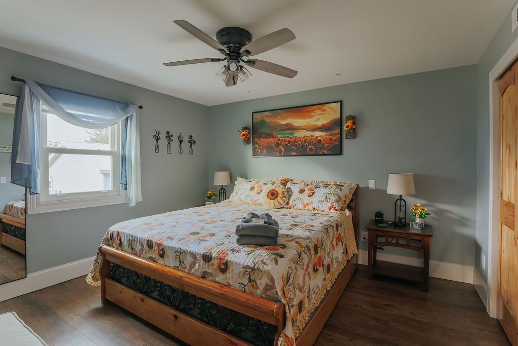 A bedroom with a bed, a ceiling fan, a painting, a nightstand, and a window with curtains.
