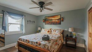 Une chambre à coucher avec un lit, un ventilateur de plafond, un tableau, une table de chevet et une fenêtre avec des rideaux.