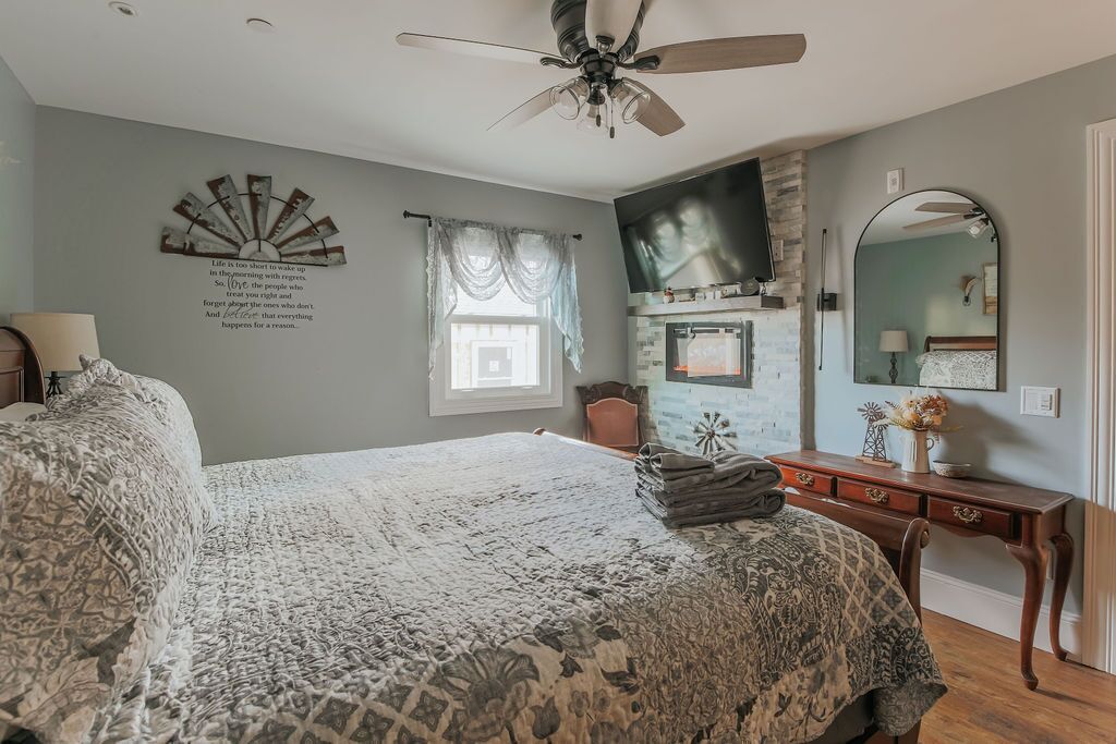 A bedroom with a large bed, a ceiling fan, a fireplace, a dresser, and a mirror.