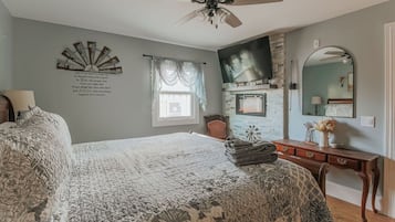 A bedroom with a large bed, a ceiling fan, a fireplace, a dresser, and a mirror.